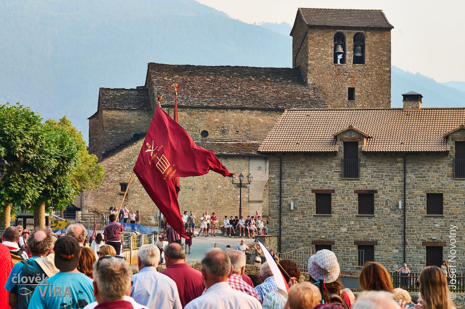 Crowd with a red flag below a stone church on a hillside